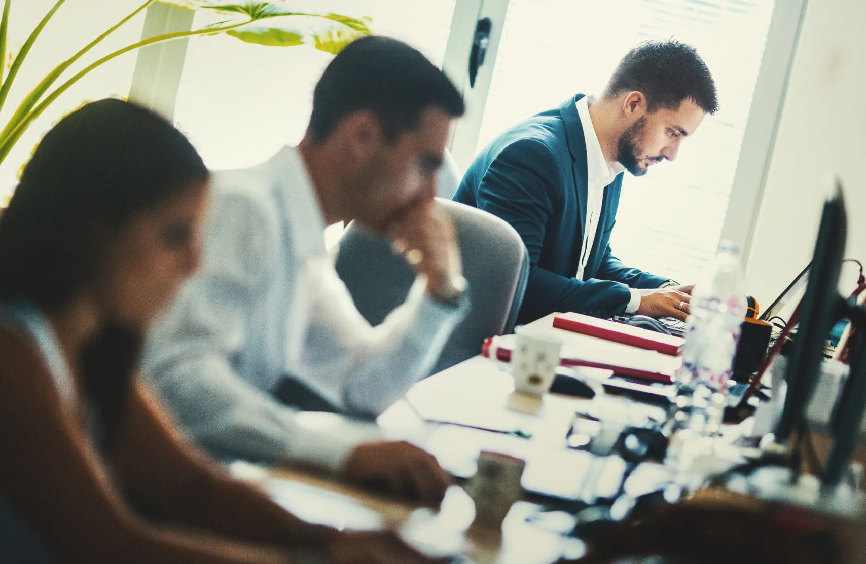 Group of mid 20's and 30's men and women engaged in application development job. They are seated by long desks back to back, each person in front of dual screen computer. Bad posture is inevitable. Back lit, toned image.